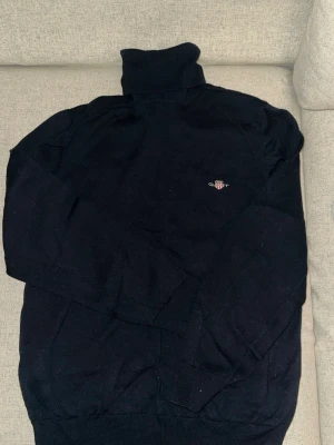 Svart polotröja från Gant - Mörkblå stickad polotröja från Gant med broderad logga på bröstet. Tröjan har lång ärm, ribbade muddar och hög polokrage. Perfekt för lager på lager och en clean look.