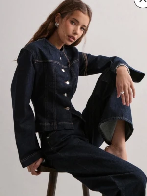 Mörkblå jeansjacka med raka ärmar - Snygg mörkblå jeansjacka i figurnära modell med raka, vida ärmar och rund halsringning. Jackan har dekorativa sömmar och knäpps med silvriga knappar framtill. Tillverkad i kraftigt denimtyg, perfekt för en trendig och avslappnad look.