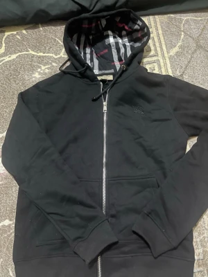Svart zip hoodie från Burberry - Snygg svart hoodie från Burberry med dragkedja framtill och klassiskt rutigt foder i huvan. Hoodien har fickor på magen och ribbade muddar vid ärmar och nederkant. Perfekt för dig som gillar stilrena plagg med exklusiva detaljer.
