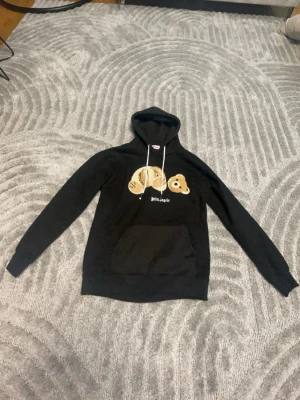 Palm Angels svart hoodie med björn - Svart hoodie från Palm Angels med stor ficka framtill och vit dragsko i huvan. På bröstet finns ett broderat motiv av en isärplockad nallebjörn och Palm Angels-loggan i vitt. Hoodien har lång ärm och avslappnad passform, perfekt för en streetwear-look.