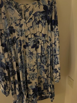 Blommig blåvit klänning med v-ringning - Säljer en luftig klänning med blått och vitt blommönster. Klänningen har v-ringning, långa ärmar och en avslappnad passform. Perfekt för dig som gillar romantiska och feminina plagg med mycket mönster.