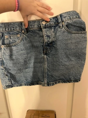 Blå jeanskjol med knappar framtill - Säljer en klassisk blå jeanskjol i kort modell med fem fickor och knappar framtill. Kjolen har en rak passform och är gjord i slitstarkt denimtyg. Perfekt till vardags för en avslappnad och trendig look.