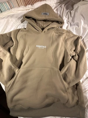 Beige Essentials Fear of God hoodie - Säljer en beige hoodie från Essentials Fear of God med stor huva och känguruficka. Hoodien har logga tryckt på bröstet och är gjord i mjukt bomullsmaterial. Perfekt för en avslappnad och trendig streetwear-look.