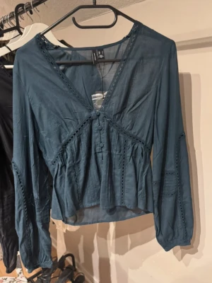 Mörkgrön blus med v-ringning Vero Moda - Trendig mörkgrön blus från Vero Moda med v-ringning, knappar framtill och dekorativa spetsdetaljer. Blusen har långa ärmar och lätt volang nedtill för en snygg siluett. Perfekt att styla med jeans eller kjol för en cool look.