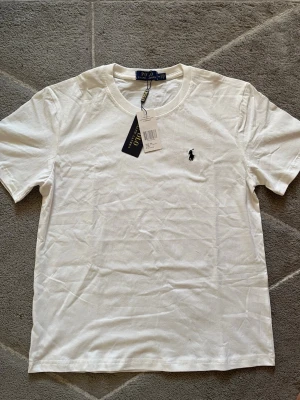 Vit t-shirt från Polo Ralph Lauren - Klassisk vit t-shirt från Polo Ralph Lauren med rund halsringning och det ikoniska broderade logotypen på bröstet. Tillverkad i mjuk bomull för en skön känsla och perfekt passform. Enkel och stilren design som funkar till allt.
