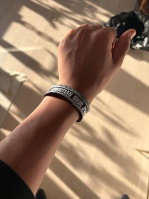 Svart Christian Dior armband med vit text och detaljer  - Snyggt svart och vitt armband från Christian Dior med tydlig logga och vävt band. Armbandet har knytning med tofsar i ändarna och passar perfekt för dig som vill ha en trendig accessoar med lyxig känsla.