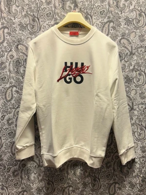 Vit sweatshirt från HUGO - Säljer en stilren vit sweatshirt från HUGO med svart och rött logotryck på bröstet. Tröjan har rund halsringning, ribbade muddar vid ärmslut och nederkant samt en relaxed passform. Perfekt för dig som gillar minimalistisk och trendig design.