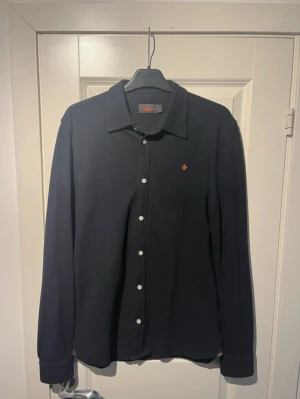  Morris skjorta   - Stilren svart skjorta från Morris med klassisk krage och vita knappar. På bröstet finns ett orange broderat emblem som ger en snygg detalj. Skjortan är långärmad och tillverkad i mjuk bomull, perfekt för en chill och clean look. 97% bomull 3% Elastan