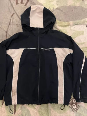 Mörkblå och vit vindjacka lets frans - Snygg blå och vit vindjacka från lets frans med huva och dragkedja framtill. Jackan har, elastiska muddar och justerbar nederkant med snörning. Perfekt för dig som gillar retrostil och sportig look.