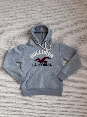 Grå Hollister hoodie med logga - Snygg grå hoodie från Hollister med stor broderad logga och texten 'California' på bröstet. Hoodien har vit dragsko i huvan och en klassisk känguruficka framtill. Tillverkad i mjukt sweatshirtmaterial, perfekt för en avslappnad stil.