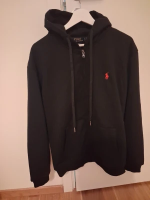 Svart hoodie från Polo Ralph Lauren - Svart hoodie från Polo Ralph Lauren med dragkedja vid halsen och klassisk röd logga broderad på bröstet. Hoodien har huva med dragsko och en stor magficka framtill. Tillverkad i mjukt bomullsmaterial, perfekt för en chill och stilren look.