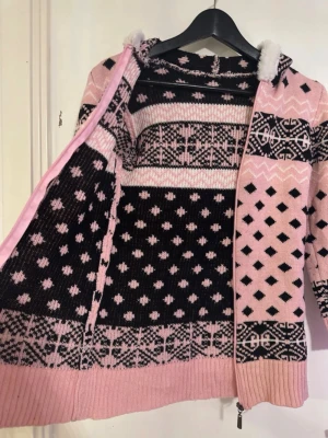 Rosa och svart stickad hoodie - Mysig stickad hoodie i rosa och svart med grafiskt mönster och dragkedja framtill. Tröjan har luva fodrad med vitt fluffigt material och ribbade muddar. Perfekt för kalla dagar när du vill vara både varm och snygg.
