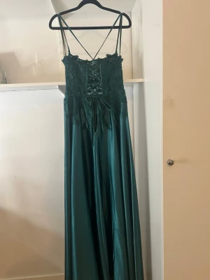 Grön långklänning med spets och snörning - Elegant långklänning i mörkgrönt satinliknande material med tunna axelband och korsad snörning i ryggen. Tyget  är dekorerat med blommig spets och små pärlor för extra detalj. Kjolen faller mjukt och ger en lyxig känsla.