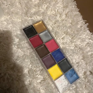 Färgstark ögonskuggspalett i plast - Säljer en ögonskuggspalett med 12 olika färger, inklusive svart, guld, silver, rosa, röd, brun, lila, blå, gul och vit. Paletten har ett genomskinligt plastlock och rektangulära pans. Perfekt för kreativa och färgglada sminkningar.