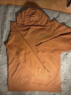 Brun hoodie från Lyle & Scott - Säljer en snygg brun hoodie från Lyle & Scott med deras klassiska gula fågel-logga på bröstet. Hoodien har huva, magficka och ribbade muddar vid ärmar och nederkant. Perfekt för en chill och avslappnad stil. Litet lagat hål på sidan men inget som syns när man bär tröjan. 