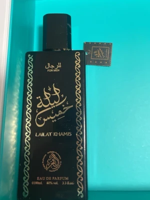 Lailat Khamis Eau de Parfum 100ml - Snygg parfymflaska i svart med gulddetaljer och orientaliskt mönster. Lailat Khamis Eau de Parfum för män, 100 ml. Flaskan har en rektangulär form och elegant design med arabiska och engelska texter. Perfekt för dig som gillar exklusiva dofter.