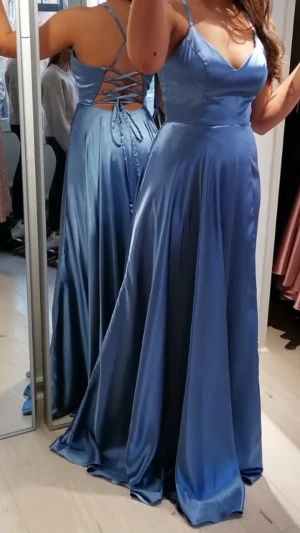 Balklänning  - Elegant blå bakkkänning i satin med v-ringad front och smala axelband. Klänningen har snörning i ryggen vilket gör att den kan justeras till många olika mått. Endast använd en kväll och tvättad på kemtvätt sen dess, nästan ings defekter, möjligtvis någon repa längst ner vid marken men inget som syns. Priset kan diskuteras. Nypris: 3300kr. 