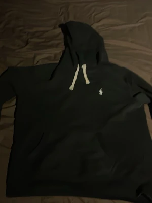 Svart hoodie från Polo Ralph Lauren - Svart hoodie från Polo Ralph Lauren med vit broderad logga på bröstet och vita dragsnören i huvan. Klassisk känguruficka framtill och mjukt material som känns skönt mot huden. Perfekt för en chill och stilren look.