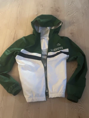 Grön och vit vindjacka från Lacoste - Snygg vindjacka från Lacoste i grönt och vitt, storlek S, startpris är 900, kan diskuteras, byxor finns att lägga till men priset höjs, fråga i DM