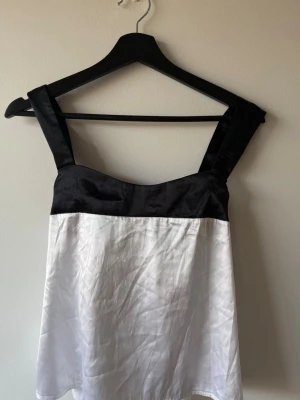 Svartvit linnetopp med breda band - Snygg svartvit linnetopp med breda axelband och knyt i ryggen. Toppen har en svart överdel och vit underdel, vilket ger en stilren kontrast. Perfekt för dig som gillar minimalistisk och trendig stil.