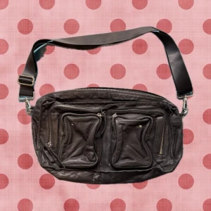 Svart axelväska i skinn med fickor - Cool svart axelväska i äkta skinn med bred justerbar axelrem. Väskan har två stora dragkedjefickor framtill och en extra ficka med dragkedja på baksidan. Perfekt för att hålla ordning på dina grejer och ge en edgy touch till din outfit.