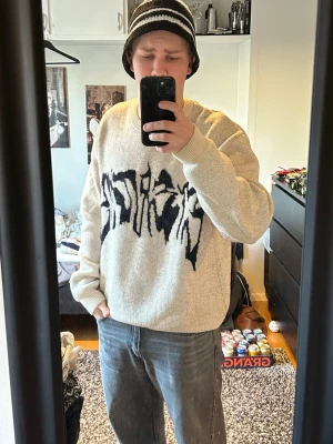 Beige stickad tröja med svart grafiskt mönster - Säljer en beige stickad tröja med ett stort svart grafiskt mönster på bröstet. Tröjan har rund halsringning och ribbstickade muddar vid ärmslut och nederkant. Perfekt för dig som gillar streetwear och unik design.