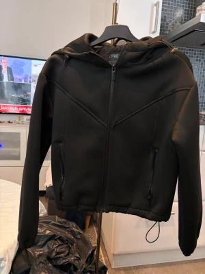 Svart hoodie med dragkedja från FEE - Svart hoodie med hel dragkedja framtill och två fickor med dragkedja. Snygga sömdetaljer över bröstet och justerbar huva. Tillverkad i ett slätt och lite tjockare material som ger en modern look.