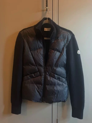 Mörkblå moncler cardigan  - Snygg mörkblå moncler cardigan med stickade ärmar och vadderad front. Jackan är i jättebra skick och knappt använd. Jackan har dragkedja framtill och Moncler-logga på ena ärmen. Perfekt för våren. 