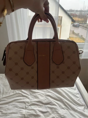 Beige och brun handväska från Catwa - Snygg handväska från Catwa i beige och brunt med två stadiga handtag. Väskan har ett diskret mönster och en bred brun panel i mitten. Tillverkad i konstläder med guldfärgad logga framtill. Perfekt för dig som gillar stilrena accessoarer.