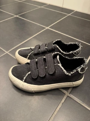 Svarta sneakers med kardborreband - Svarta sneakers från Even&Odd i denimliknande tyg med råa, fransiga kanter och tre breda kardborreband. Skorna har vit sula och en cool, avslappnad look som sticker ut. Perfekta för dig som gillar streetstyle och vill ha något unikt på fötterna.