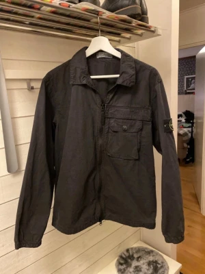 Svart overshirt från Stone Island - Snygg svart overshirt från Stone Island med dragkedja framtill, klassisk krage och en bröstficka med knapp. Ikoniska Stone Island-märket sitter på vänster ärm. Tillverkad i ett lätt bomullsmaterial, perfekt för lager på lager och streetwear-stil.