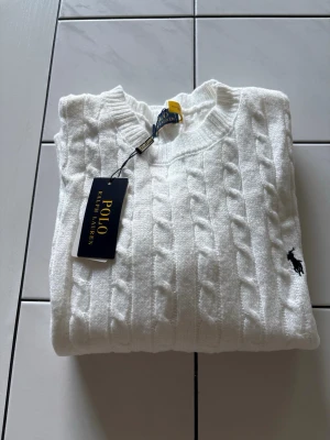 Vit kabelstickad tröja Polo Ralph Lauren - Vit kabelstickad tröja från Polo Ralph Lauren med rund halsringning och klassisk svart broderad logga på bröstet. Tröjan har ribbade muddar och är tillverkad i ett mjukt, värmande material. Perfekt för en stilren och tidlös look.