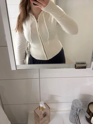 Vit stickad hoodie med dragkedja från Zara - Snygg vit stickad hoodie med dragkedja framtill och dragsko i huvan. Modellen är figurnära med ribbade muddar och midja. Perfekt för en chill och stilren look. Materialet känns mjukt och bekvämt mot huden.