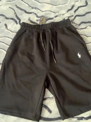 Svarta och vita shorts från Polo Ralph Lauren - Två par shorts från Polo Ralph Lauren, ett par svarta och ett par vita. Båda har elastisk midja med snörning och klassisk Polo-logga broderad på vänster ben. Perfekta för en avslappnad stil och tillverkade i mjuk bomull.