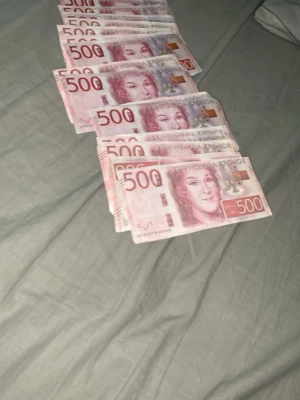 Svenska 500-kronors sedlar som är fejk som kan vara kul att flexa me - Ett antal svenska 500-kronors sedlar i röd och rosa nyans med porträtt och säkerhetsdetaljer. Sedlarna är placerade på en grå yta och har tydliga markeringar och hologram. Perfekt för samlare eller som rekvisita. Men dom är fejk