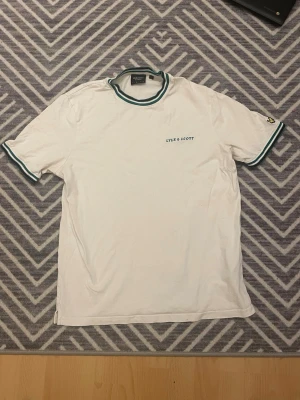 Vit t-shirt från Lyle & Scott - Vit t-shirt från Lyle & Scott med gröna och vita ränder på krage och ärmslut. Klassisk passform och korta ärmar. Diskret logga på bröstet och liten broderad detalj på ena ärmen. Perfekt till jeans eller shorts.