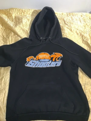 Svart Trapstar dress - Svart hoodie från Trapstar med stor färgglad 'Trapstar Shooters'-logga i orange, blått och vitt på bröste/byxorna. Klassisk känguruficka och justerbar huva med snören. Perfekt för dig som gillar streetwear och vill sticka ut med en fet logga. Enda som fattas är snöret till byxorna!