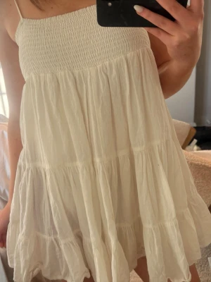 Vit somrig kortklänning med volang - Luftig vit kortklänning med smock upptill och tunna axelband. Klänningen har flera volanglager och är tillverkad i ett lätt, skrynkligt tyg som ger en avslappnad känsla. Perfekt för varma dagar och enkel att styla.