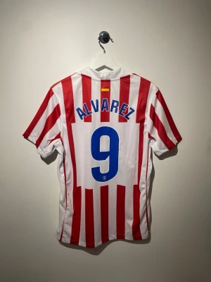 Atletico Madrid matchtröja Nike röd/vit - Snygg Atletico Madrid matchtröja med röda och vita ränder, blå Nike-logga och klubbmärke på bröstet. Nummer 9 och namnet Alvarez på ryggen. Sponsorlogga Riyadh Air framtill. Perfekt för fotbollsfans!