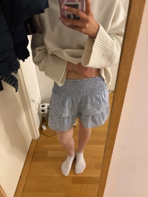 Blå blommig volangkjol - Supersöt kort blå kjol med vitt blommönster och volangdetaljer. Kjolen har en smockad midja som gör den bekväm och lätt att bära. Perfekt för vår och sommar, och passar till både stickad tröja och t-shirt.   