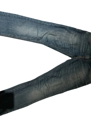 Blå jeans från Nudie Jeans - Snygga blå jeans från Nudie Jeans med klassisk femficksdesign och orangea sömmar. Jeansen har en något tvättad look och raka ben, samt slitna detaljer vid benslut. Perfekta för en avslappnad stil.