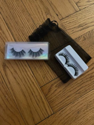 Svarta lösögonfransar i två förpackningar - Två par svarta lösögonfransar i olika stilar, levereras i plastförpackningar. Ena asken har holografisk effekt och den andra är vit. Fransarna är täta och voluminösa, perfekta för att framhäva ögonen. Medföljer gör en svart nätpåse för förvaring.