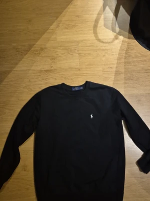 Svart tröja från Polo Ralph Lauren - Svart långärmad tröja från Polo Ralph Lauren med klassisk rund halsringning och den ikoniska vita logotypen broderad på bröstet. Enkel och stilren design som passar perfekt till jeans eller chinos.