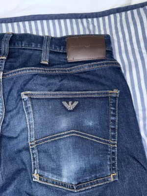 Mörkblå jeans från Emporio Armani - Snygga mörkblå jeans från Emporio Armani med klassisk femficksdesign och logga på bakfickan. Jeansen har raka ben och kontrastsömmar. Perfekta för dig som gillar stilrena och tidlösa plagg. Materialet är denim i bomull. Knappen är lite lös går att fixa själv om vet hur eller hos en skräddare, inget som hindrar den från att stängas iallafall.