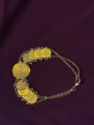 Guldarmband med myntberlocker - Snyggt armband i guldfärgad metall med flera runda myntberlocker i olika storlekar. Mynten har detaljerade mönster och armbandet har en tunn kedja med karbinlås. Perfekt accessoar för dig som gillar unika smycken med boho-vibe.