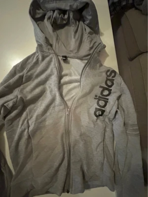 Grå hoodie med dragkedja från Adidas - Snygg grå hoodie från Adidas med dragkedja framtill och stor huva. Adidas-loggan i svart tryck längs ena sidan ger en sportig vibe. Tillverkad i mjukt material som känns skönt mot huden. Perfekt för chill eller träning.