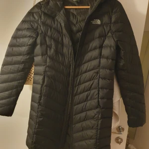 Svart lång dunjacka The North Face - Säljer en svart lång tunn vår dunjacka/kappa från The North Face med huva och dragkedja framtill. Jackan har quiltad design och logga på bröstet och ryggen. Fint och bra skick,använd fåtal gånger dock några klösmärken från mina katter, se bilder! 