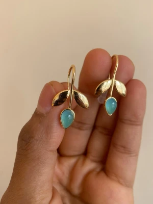 Guldörhängen med blå pärldetalj - Unika örhängen i guldfärgad metall med två blanka blad och en droppformad blå pärldetalj längst ner. Snygg och stilren design som sticker ut med sin kombination av guld och blått. Perfekta för dig som gillar naturinspirerade accessoarer. material: steel