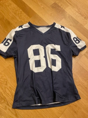 Blå sportig t-shirt med nummer 86 - Mörkblå t-shirt med vita detaljer och stort tryck av siffran 86 på bröstet och ärmarna. V-ringad modell med korta ärmar och vita stjärnor på axlarna. 