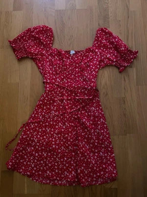 Röd blommig omlottklänning - Söt röd omlottklänning med vita blommor och kort puffärm. Klänningen har knyt i midjan och.